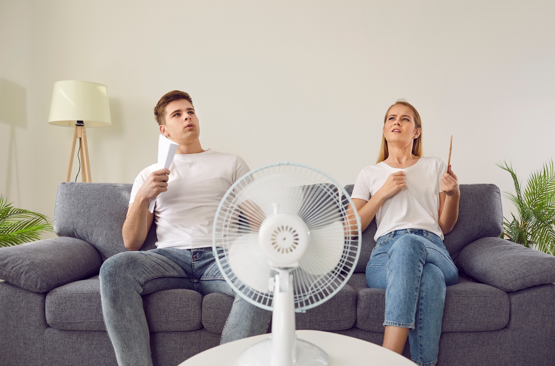 A man and woman sitting on couch in hot living room, fanning themselves with papers.