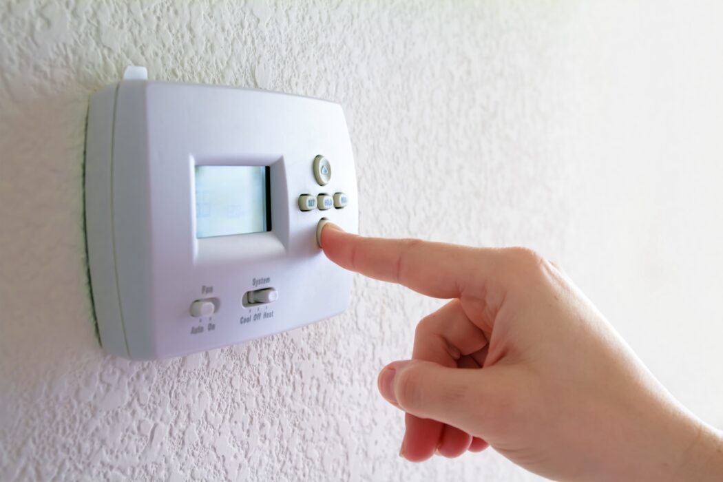 A hand adjusts the temperature on a wall-mounted digital thermostat, addressing HVAC issues on a textured white wall.
